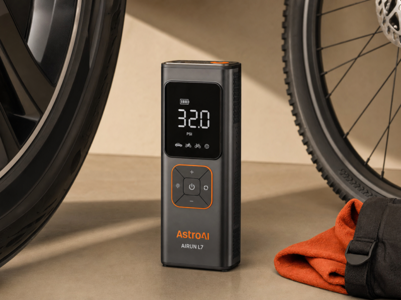 AstroAI AIRUN L7 cordless tire inflator standing upright between a car wheel and a bicycle wheel on a warm neutral background